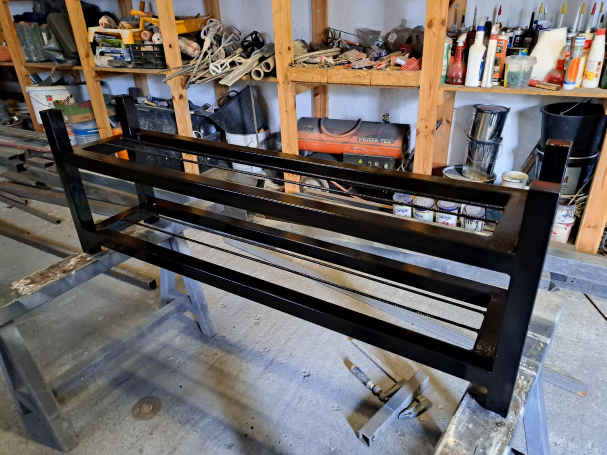 Czarna, metalowa konstrukcja z profili na stojakach warsztatowych. Widoczne spawy i elementy metalowe. Tło warsztatowe, regały z narzędziami i farbami.