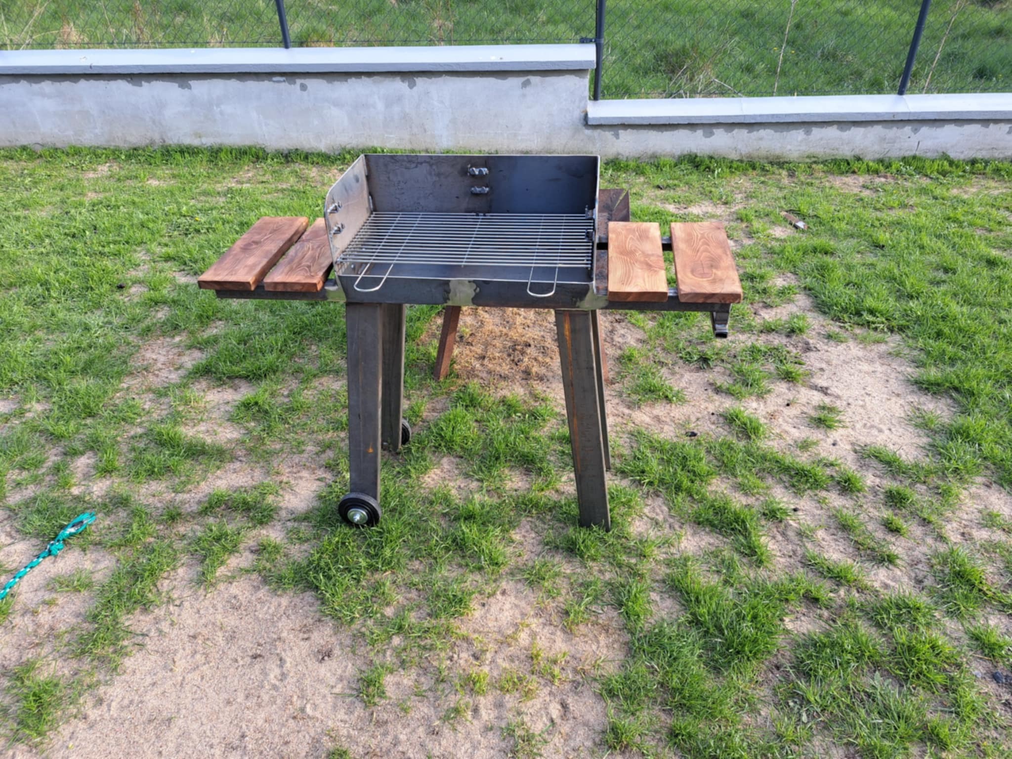 Metalowy grill ogrodowy na kółkach z drewnianymi półkami, ustawiony na trawie przed betonowym ogrodzeniem. Widoczne spawane elementy konstrukcyjne.