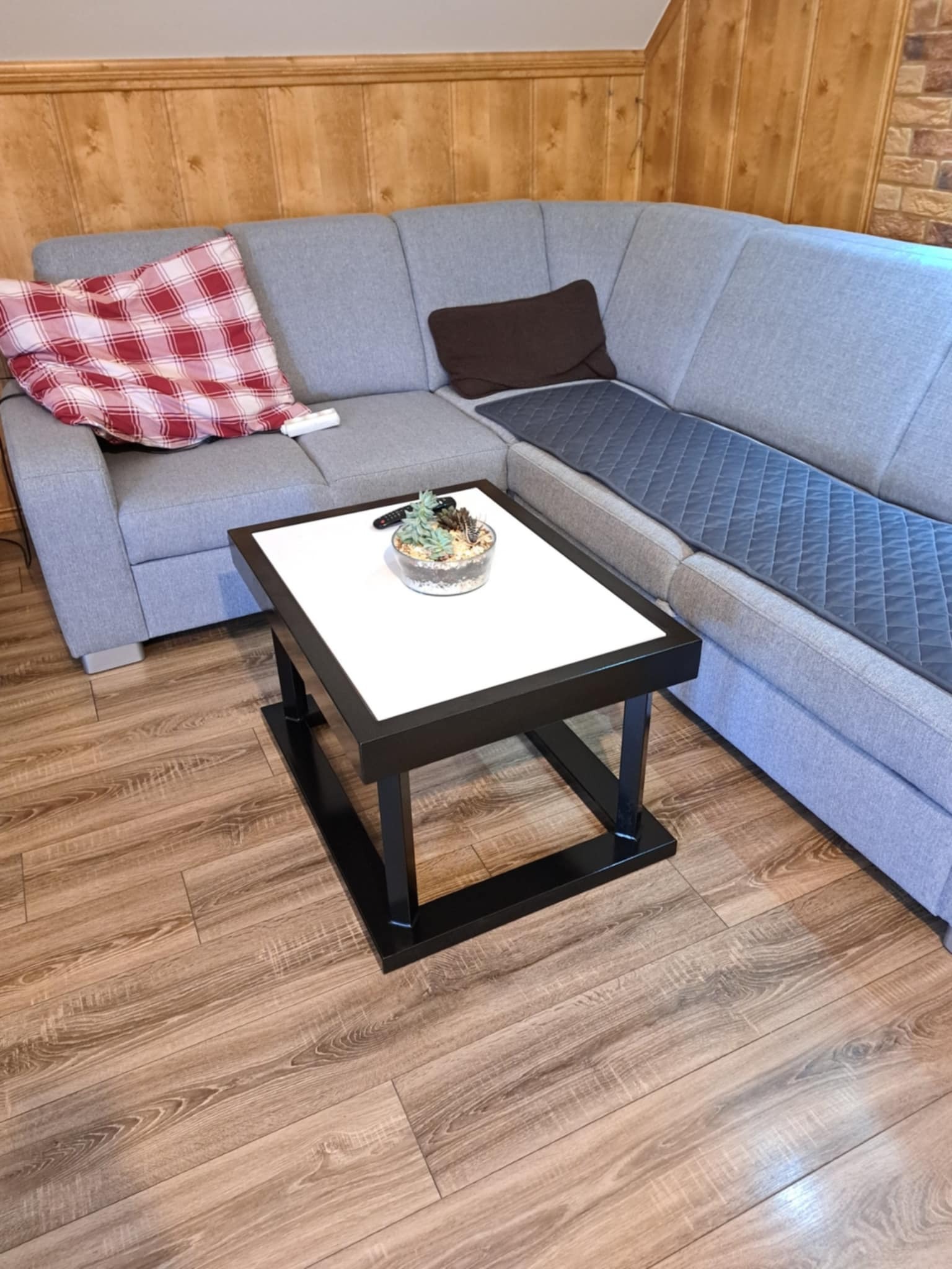 Nowoczesny stolik kawowy z białym blatem i czarną ramą, na drewnianej podłodze, obok szarego narożnika z poduszkami. Wazonik z sukulentami na blacie.
