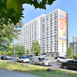 Alicja Dudewicz ALKAVALUE - Blok mieszkalny z billboardem w tle, widziany z perspektywy ulicy z przejeżdżającymi samochodami i rowerzystą na chodniku w słoneczny dzień. Zieleń drzew w kadrze.