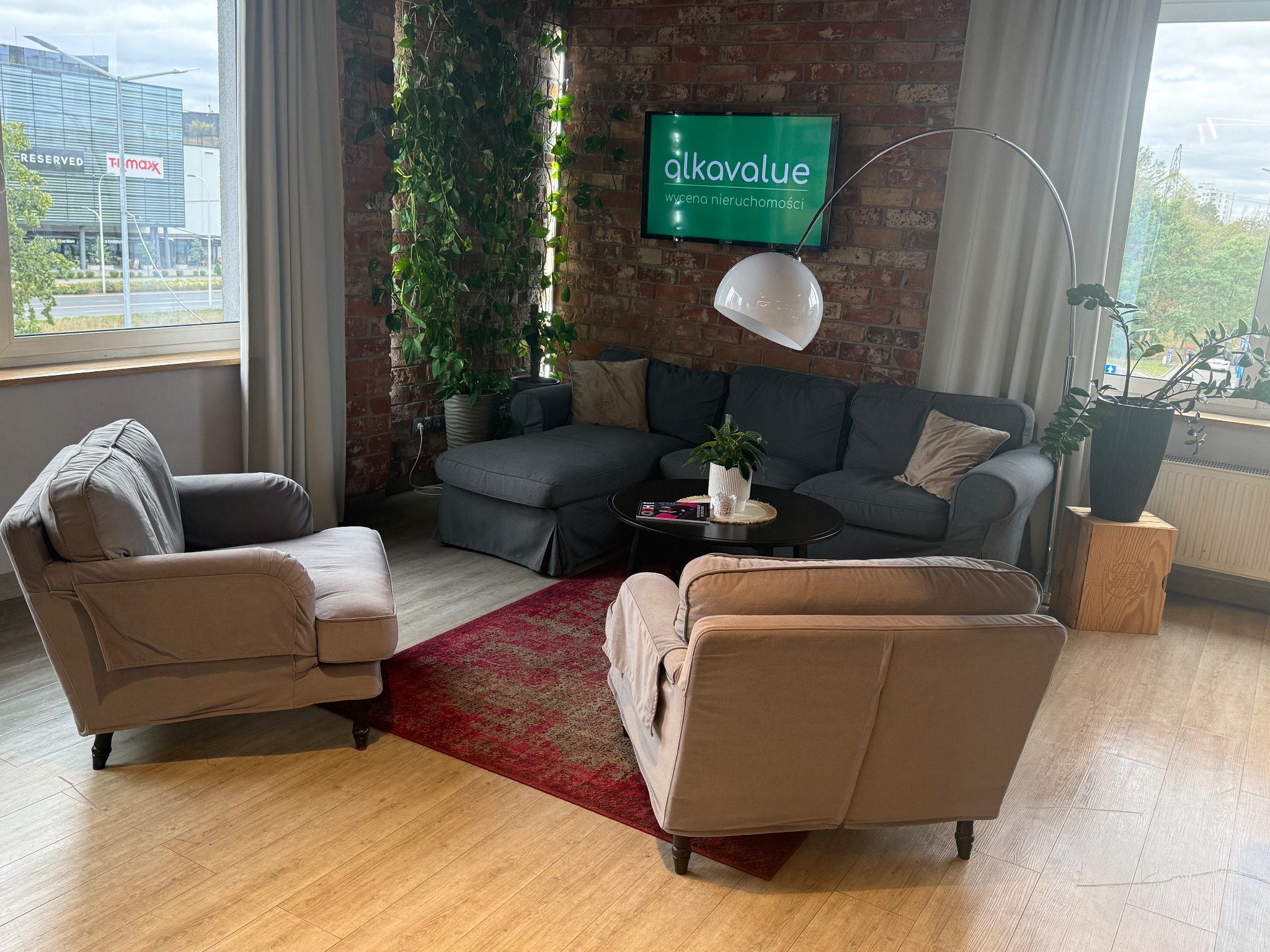 Stylowe wnętrze biura z ceglaną ścianą, szarą sofą, fotelami i roślinami. Na ścianie telewizor z logo firmy 'alkavalue wycena nieruchomości'.