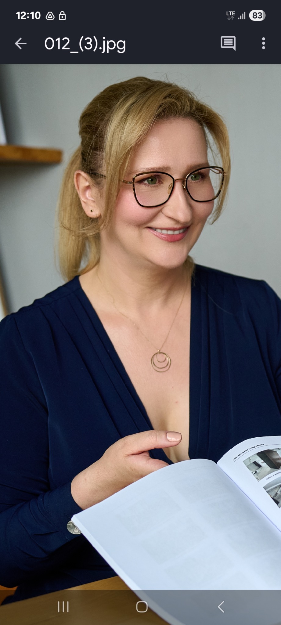 Kobieta w okularach, z uśmiechem, przegląda dokumentację fotograficzną wnętrz. Widoczny złoty naszyjnik i granatowa bluzka.