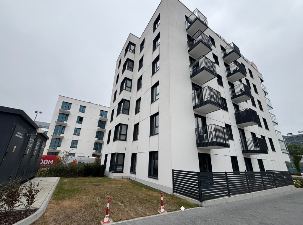 Nowoczesny budynek mieszkalny z balkonami, biała elewacja z czarnymi ramami okiennymi i balustradami. Widok z dołu, pochmurne niebo, fragment trawnika i chodnika.