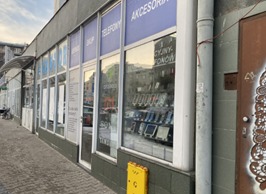 Fasada sklepu z telefonami i akcesoriami, widoczna wystawa z ekspozycją produktów. Szary budynek z rurą spustową i graffiti po prawej stronie. Ulica w tle.