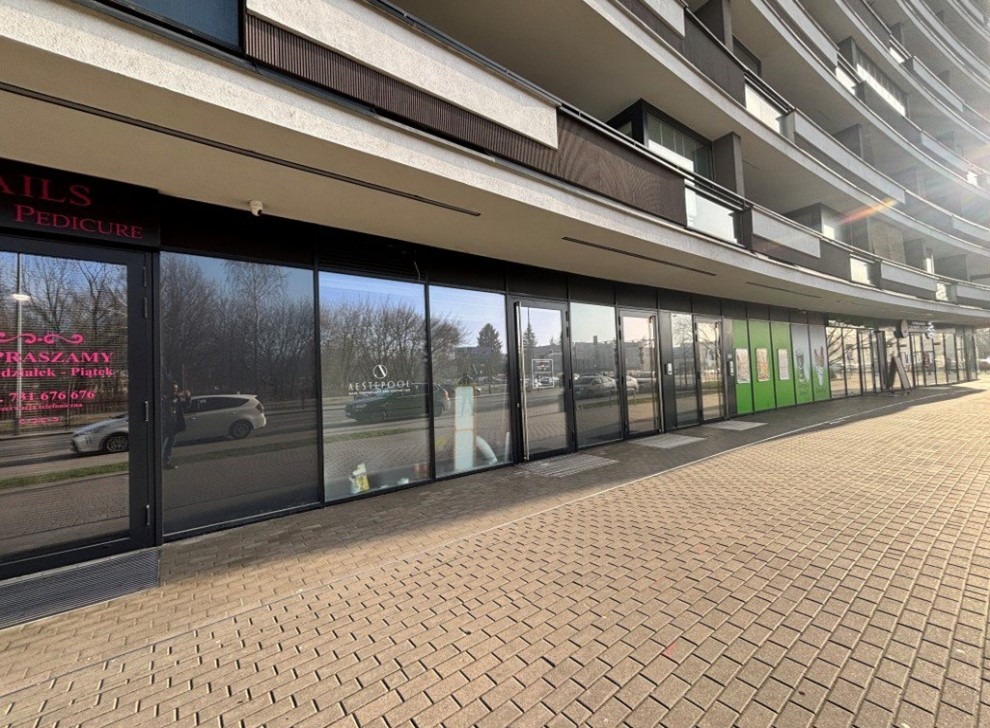 Fasada budynku z lokalami usługowymi na parterze, widoczne witryny sklepowe i biurowe, chodnik z kostki brukowej, budynek z balkonami w tle, słoneczny dzień.