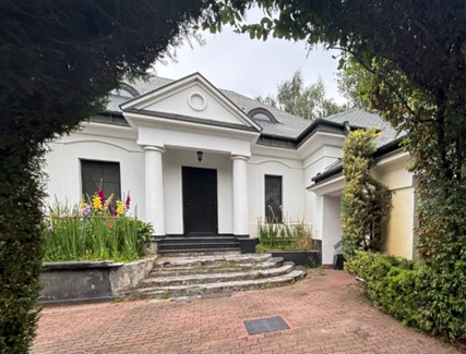 Elegancki, biały dom z kolumnami i ciemnym dachem, otoczony zielenią. Uwagę przyciągają kolorowe kwiaty przed wejściem i brukowa ścieżka prowadząca do budynku.