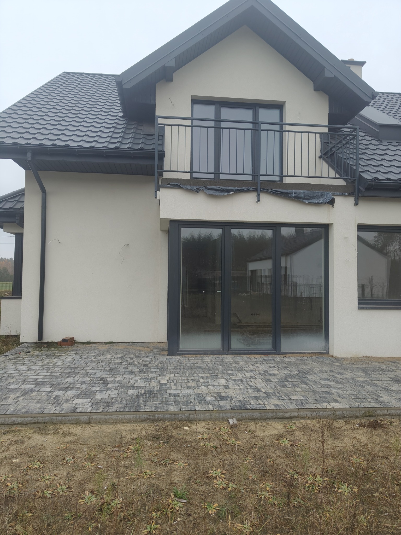 Nowoczesny dom jednorodzinny z szarą dachówką i dużymi oknami, widok z zewnątrz na świeżo wyłożony taras z kostki brukowej. Częściowo wykończony ogród.
