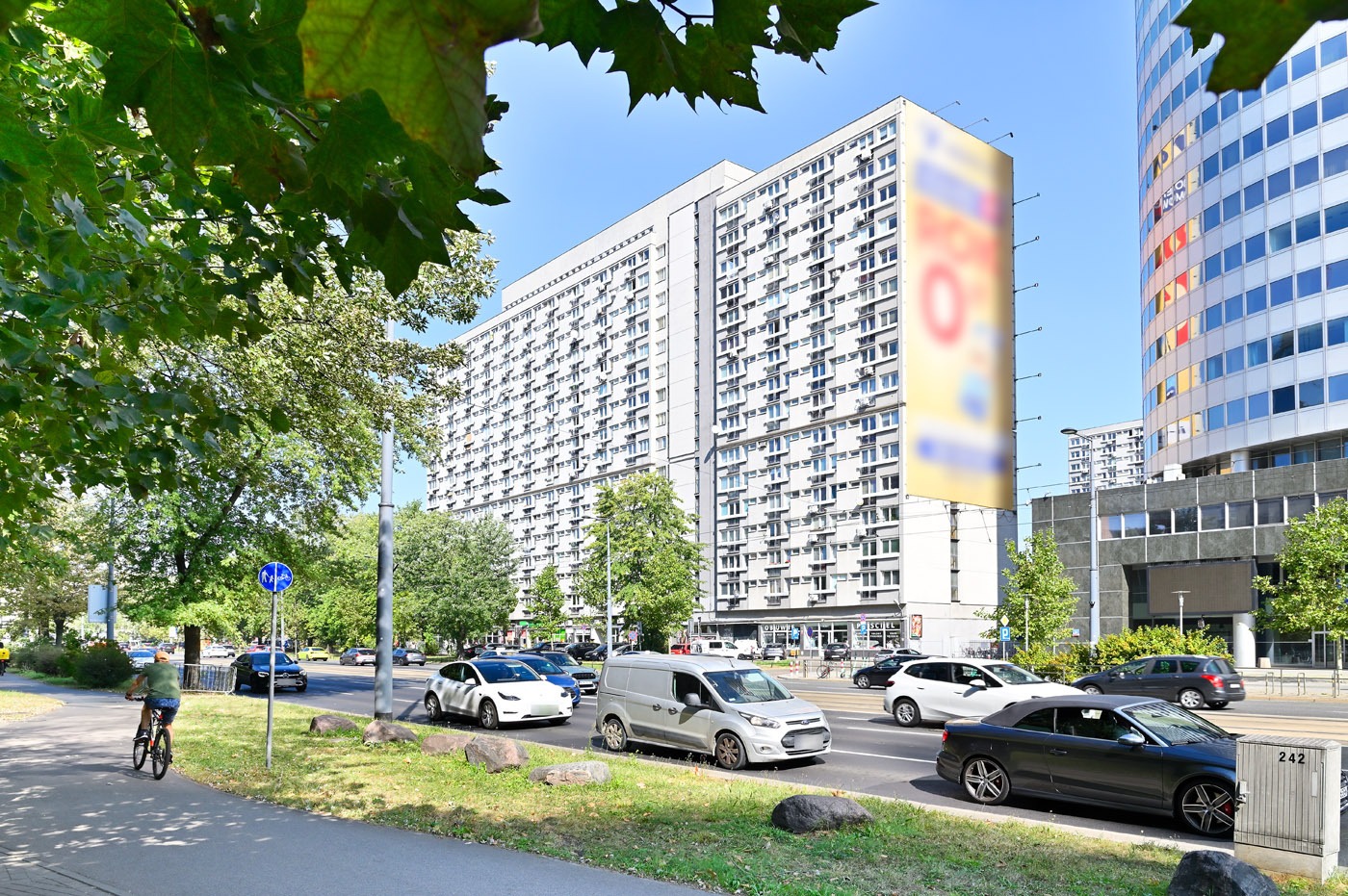 Blok mieszkalny z billboardem w tle, widziany z perspektywy ulicy z przejeżdżającymi samochodami i rowerzystą na chodniku w słoneczny dzień. Zieleń drzew w kadrze.