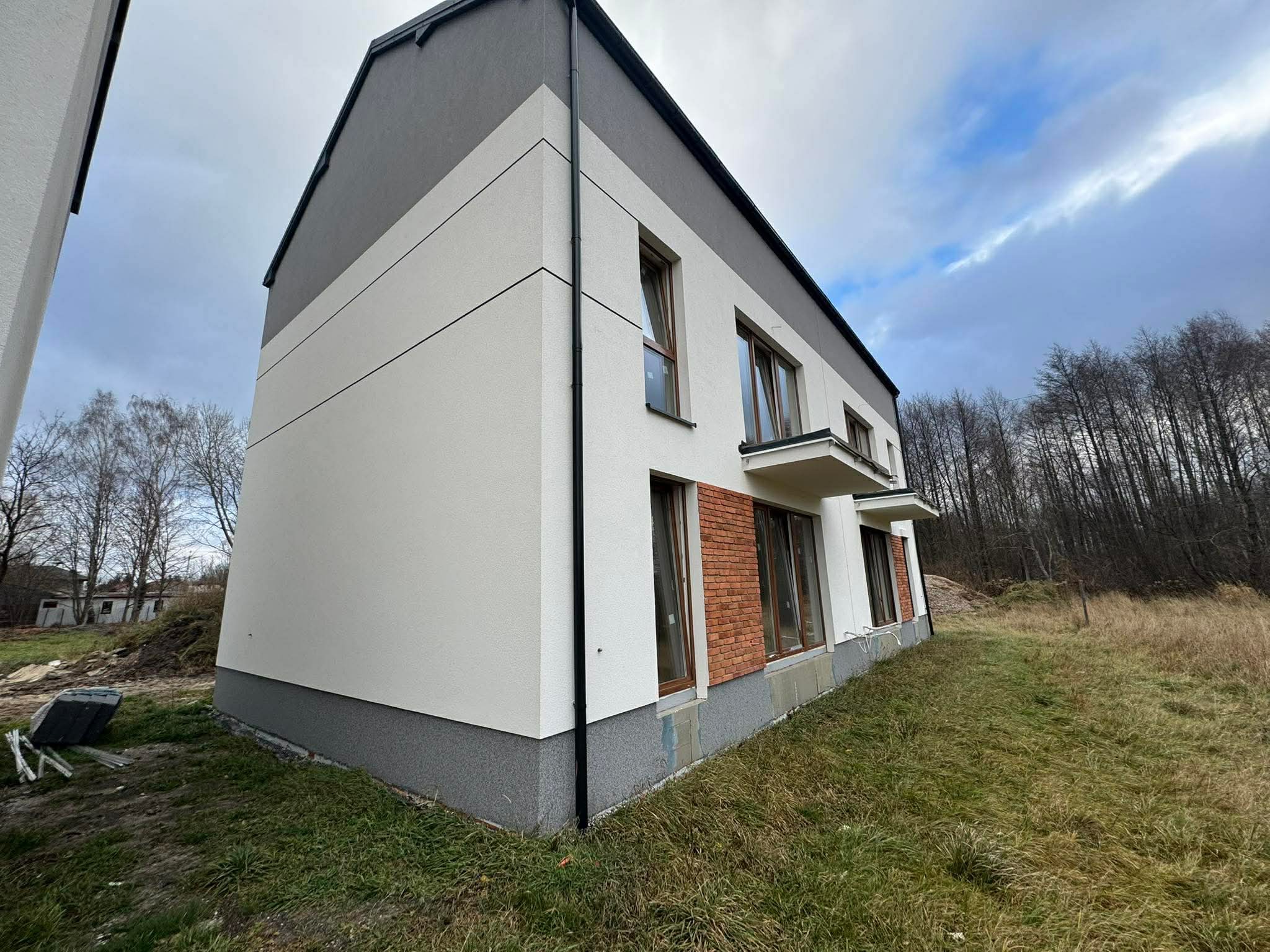 Nowoczesny, trzypiętrowy dom z szarą elewacją i ceglanym akcentem, widziany z niskiego kąta, na tle pochmurnego nieba i drzew. Widoczny fragment trawnika.