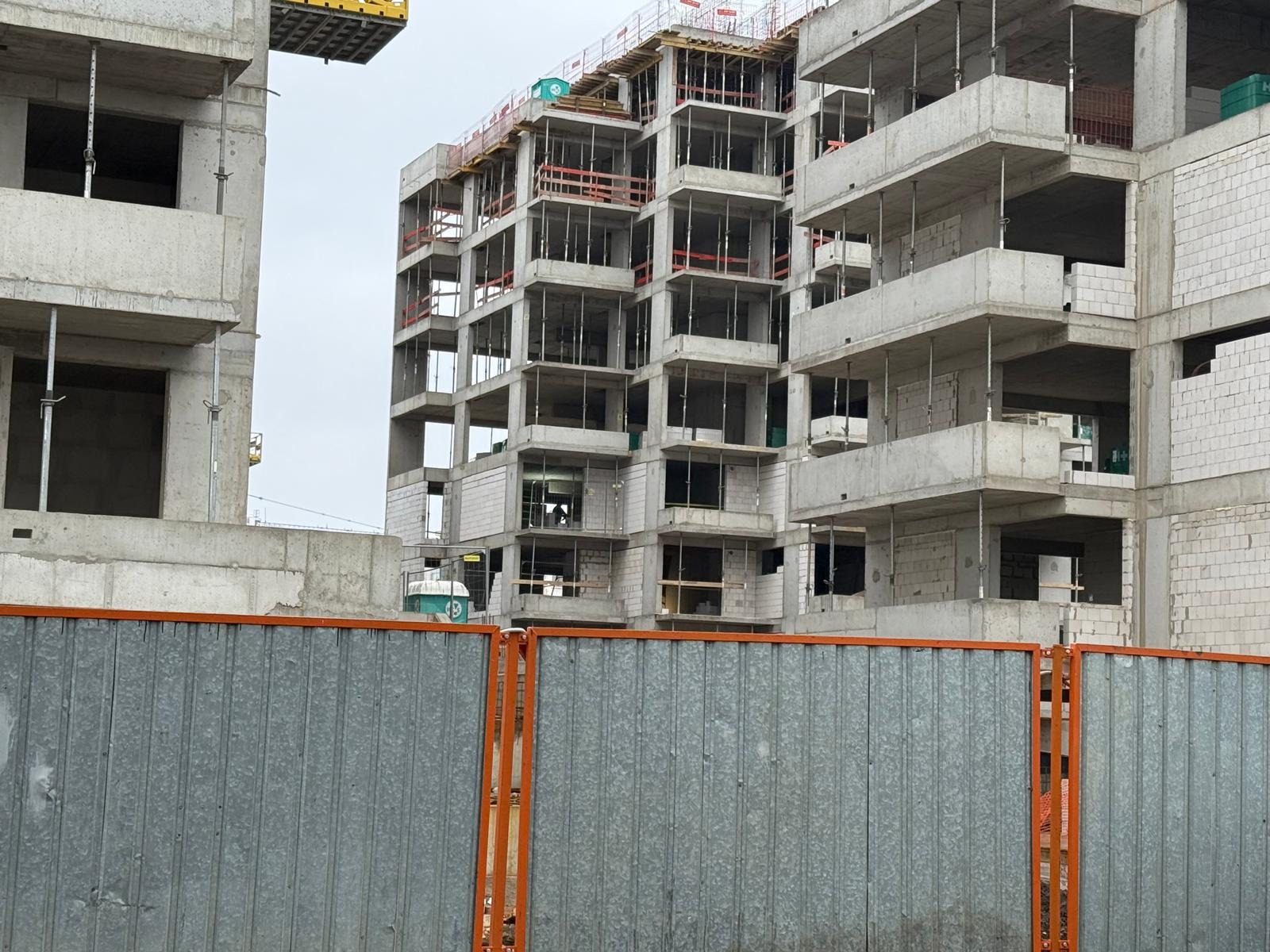 Surowy budynek w trakcie budowy, widoczny przez szarą metalową barierkę z pomarańczowymi elementami. Konstrukcja z betonu i pustaków, wielopiętrowa, w tle pochmurne niebo.