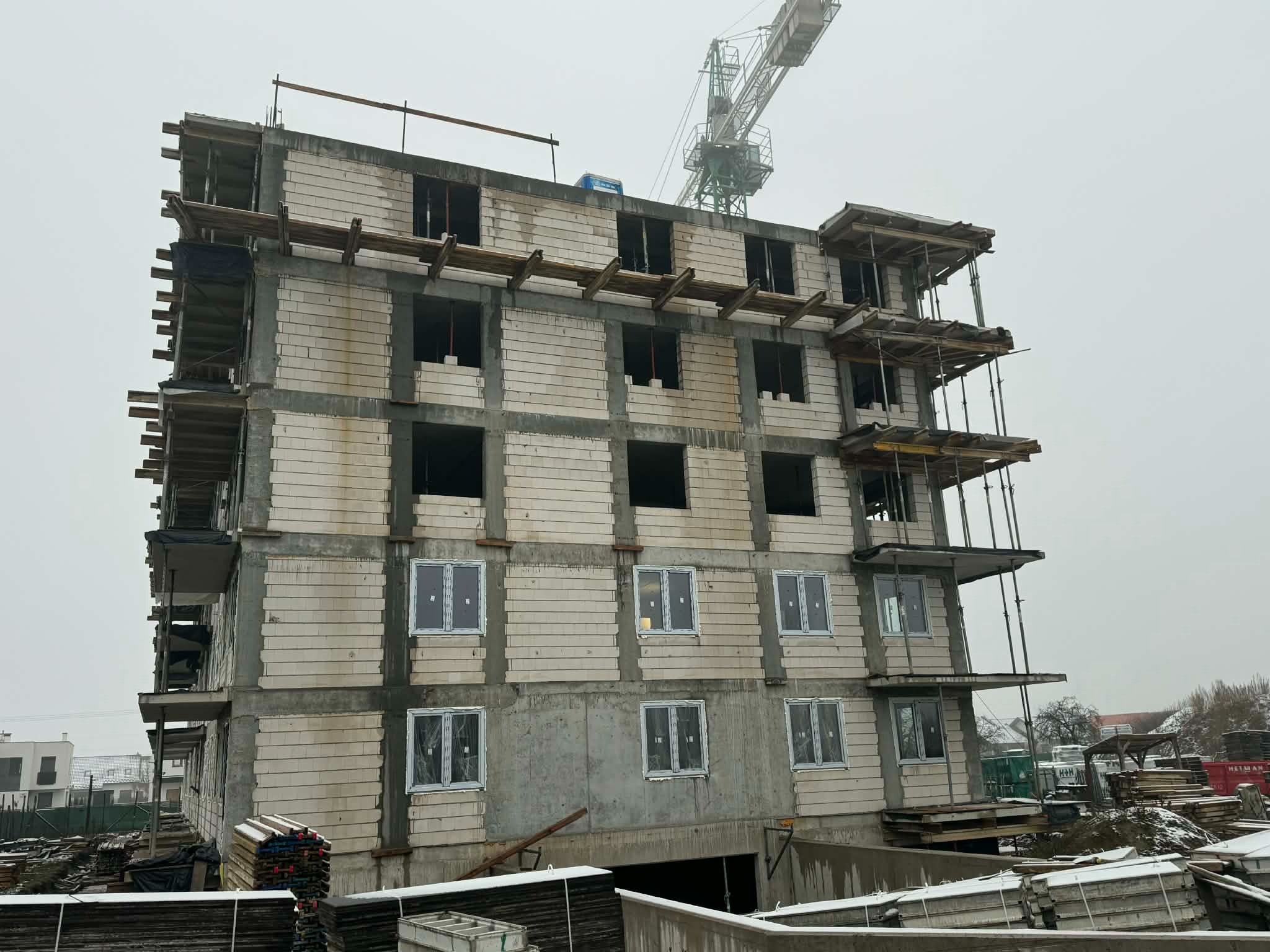 Wznoszony budynek wielopiętrowy z białego bloczka, z widocznymi oknami na niższych kondygnacjach i rusztowaniami. W tle żuraw budowlany i pochmurne niebo.