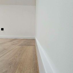 Rem-Perfect - Jasny parkiet ułożony przy białej ścianie z listwą przypodłogową. Widoczny fragment gniazdka elektrycznego. Minimalistyczne wykończenie wnętrza.