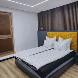 Rem-Perfect - Nowoczesna sypialnia z drewnianą ścianą, żółtym zagłówkiem i podświetlanym sufitem. Parkiet na podłodze i okno z żaluzjami dodają przytulności.