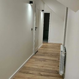 Rem-Perfect - Biały korytarz na poddaszu z drewnianą podłogą, grzejnikiem i czarnymi lampami ściennymi. Widoczne drzwi i skos dachu.