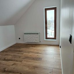 Rem-Perfect - Jasne poddasze z drewnopodobną podłogą, białymi ścianami i oknem z widokiem na zimowy krajobraz. Wisząca żarówka dodaje surowego charakteru.