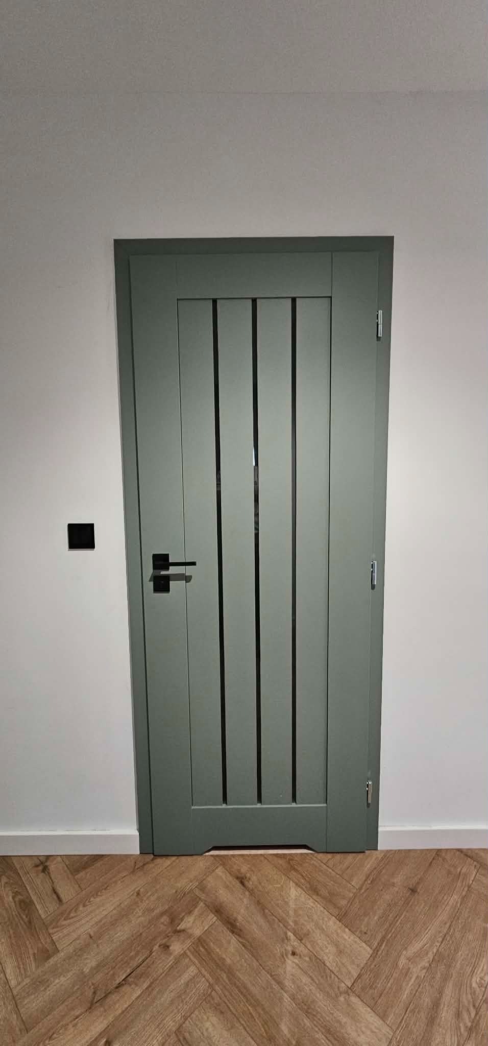 Zamknięte, szare drzwi wewnętrzne z czarną klamką i trzema pionowymi przeszkleniami, na tle białej ściany i podłogi z paneli ułożonych w jodełkę.