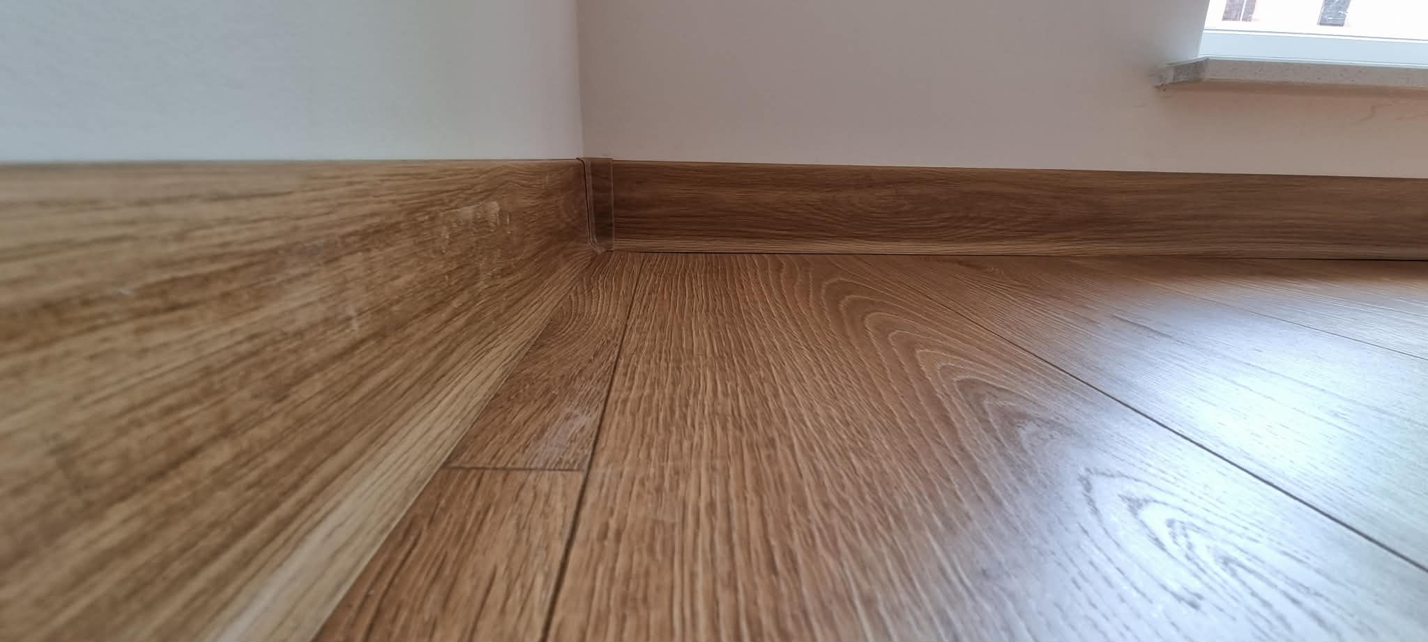 Ukończona podłoga z paneli w odcieniach brązu, z dopasowaną listwą przypodłogową. Widoczny fragment okna z parapetem. Ujęcie z niskiej perspektywy.
