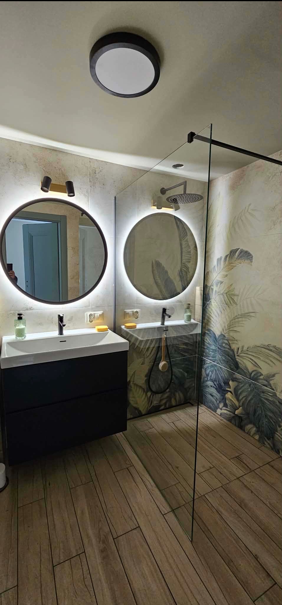 Nowoczesna łazienka z drewnopodobną podłogą, okrągłymi lustrami z podświetleniem LED i kabiną prysznicową typu walk-in z motywem tropikalnych liści na ścianie.