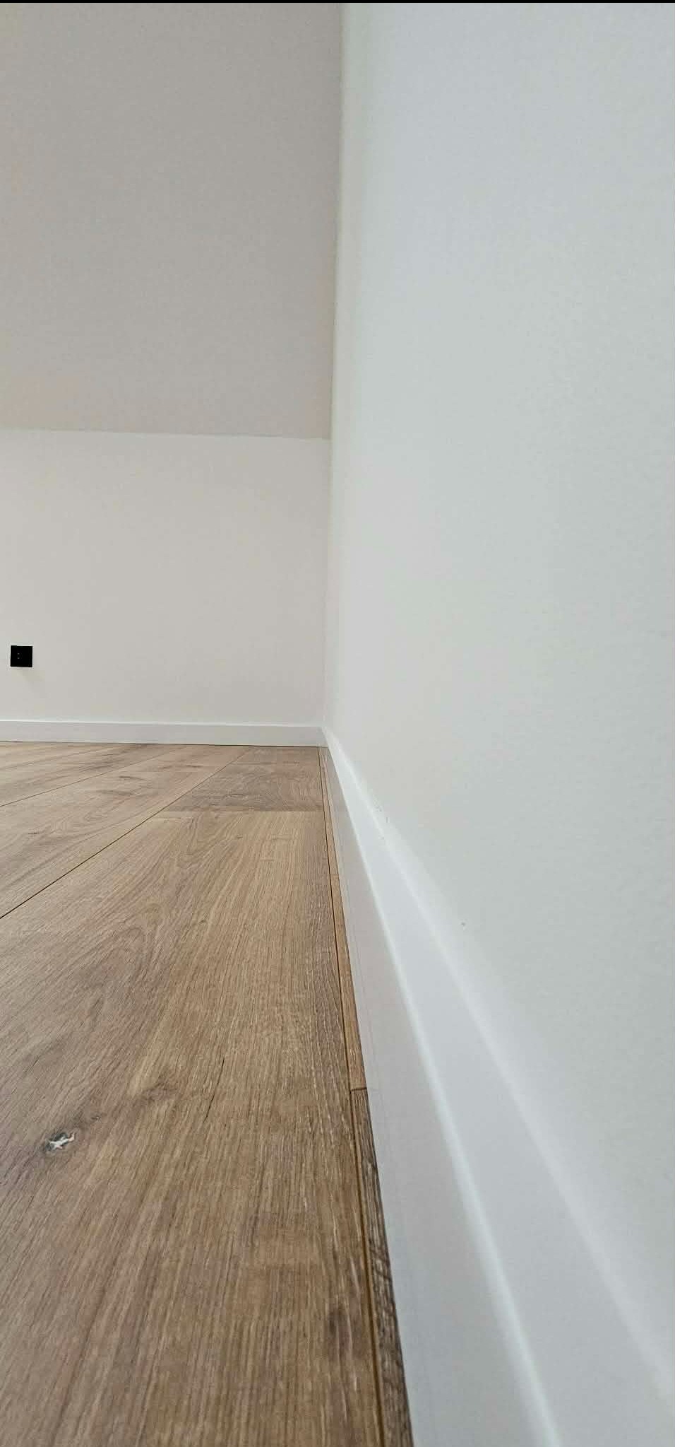Jasny parkiet ułożony przy białej ścianie z listwą przypodłogową. Widoczny fragment gniazdka elektrycznego. Minimalistyczne wykończenie wnętrza.