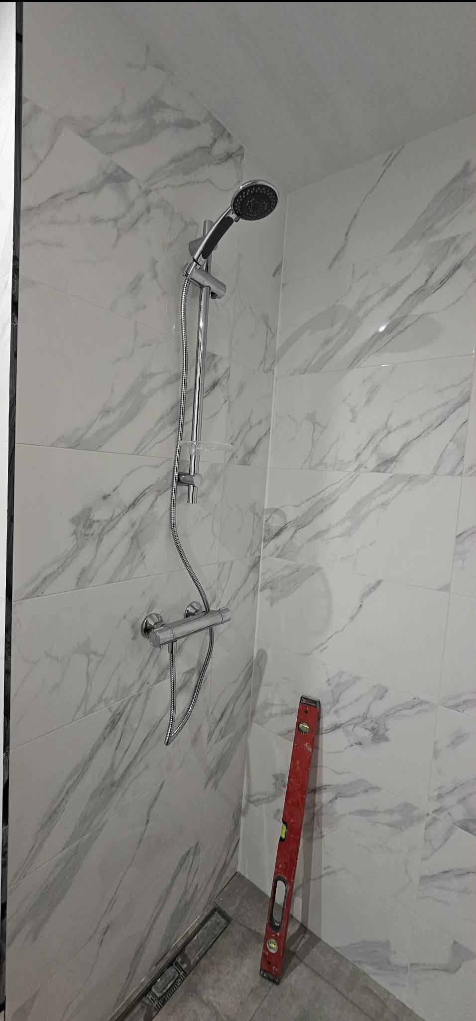 Wykończona kabina prysznicowa z marmurowymi płytkami, chromowaną armaturą i czerwoną poziomicą. Widoczny odpływ liniowy w podłodze.