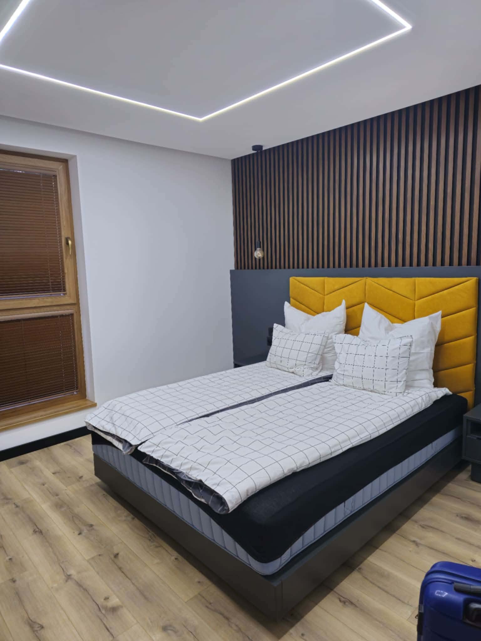 Nowoczesna sypialnia z drewnianą ścianą, żółtym zagłówkiem i podświetlanym sufitem. Parkiet na podłodze i okno z żaluzjami dodają przytulności.