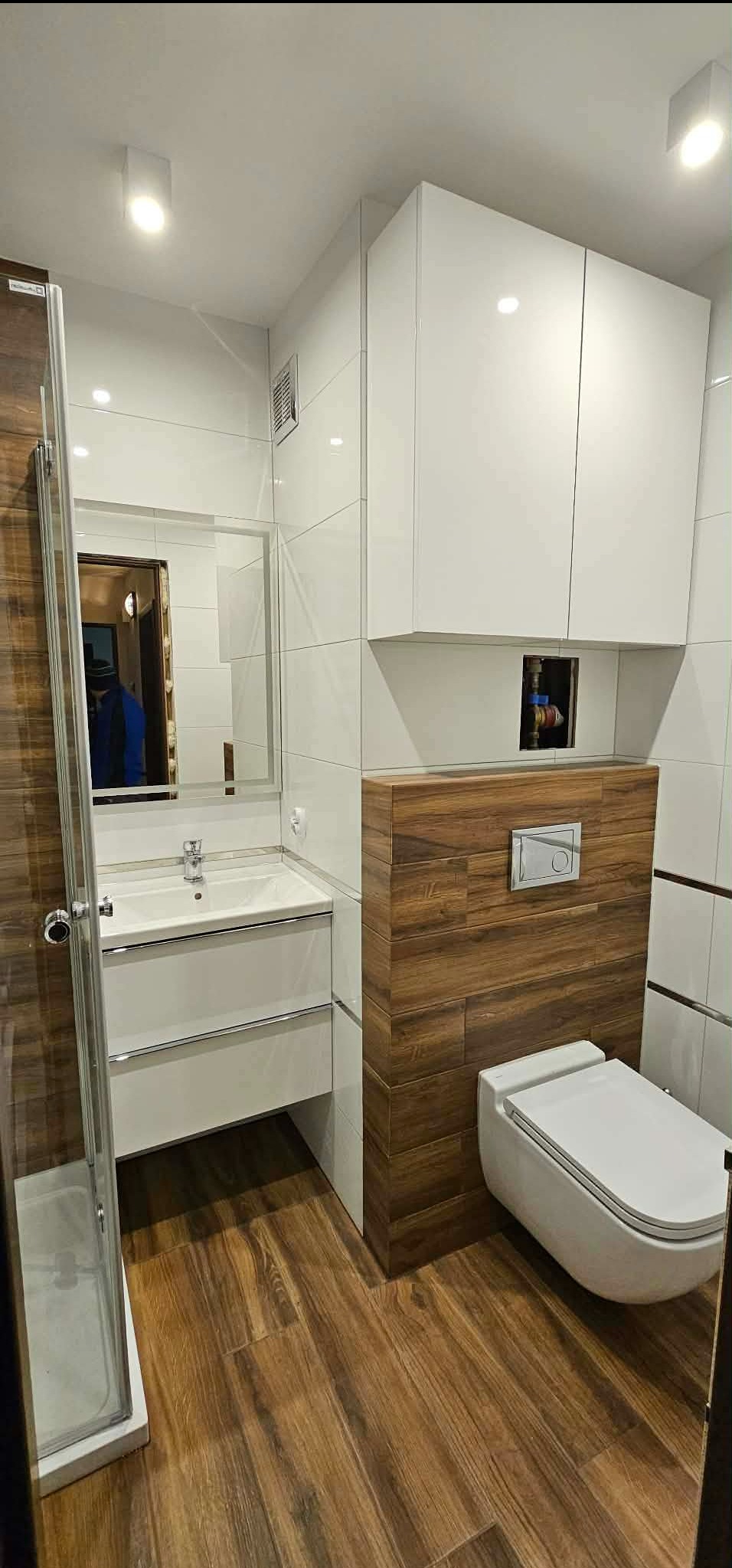 Nowoczesna łazienka po remoncie: białe płytki, drewnopodobne panele, podwieszana toaleta i szafka z umywalką. Lustro odbija fragment wnętrza.