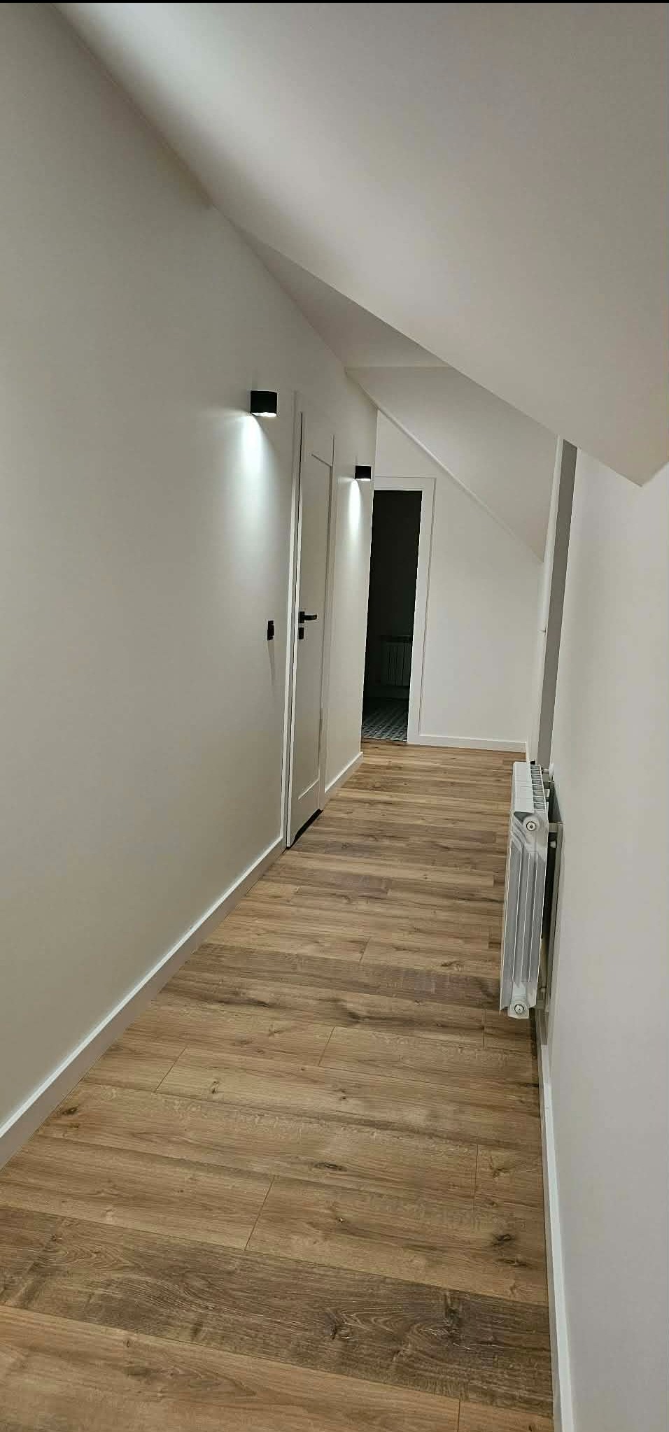 Biały korytarz na poddaszu z drewnianą podłogą, grzejnikiem i czarnymi lampami ściennymi. Widoczne drzwi i skos dachu.