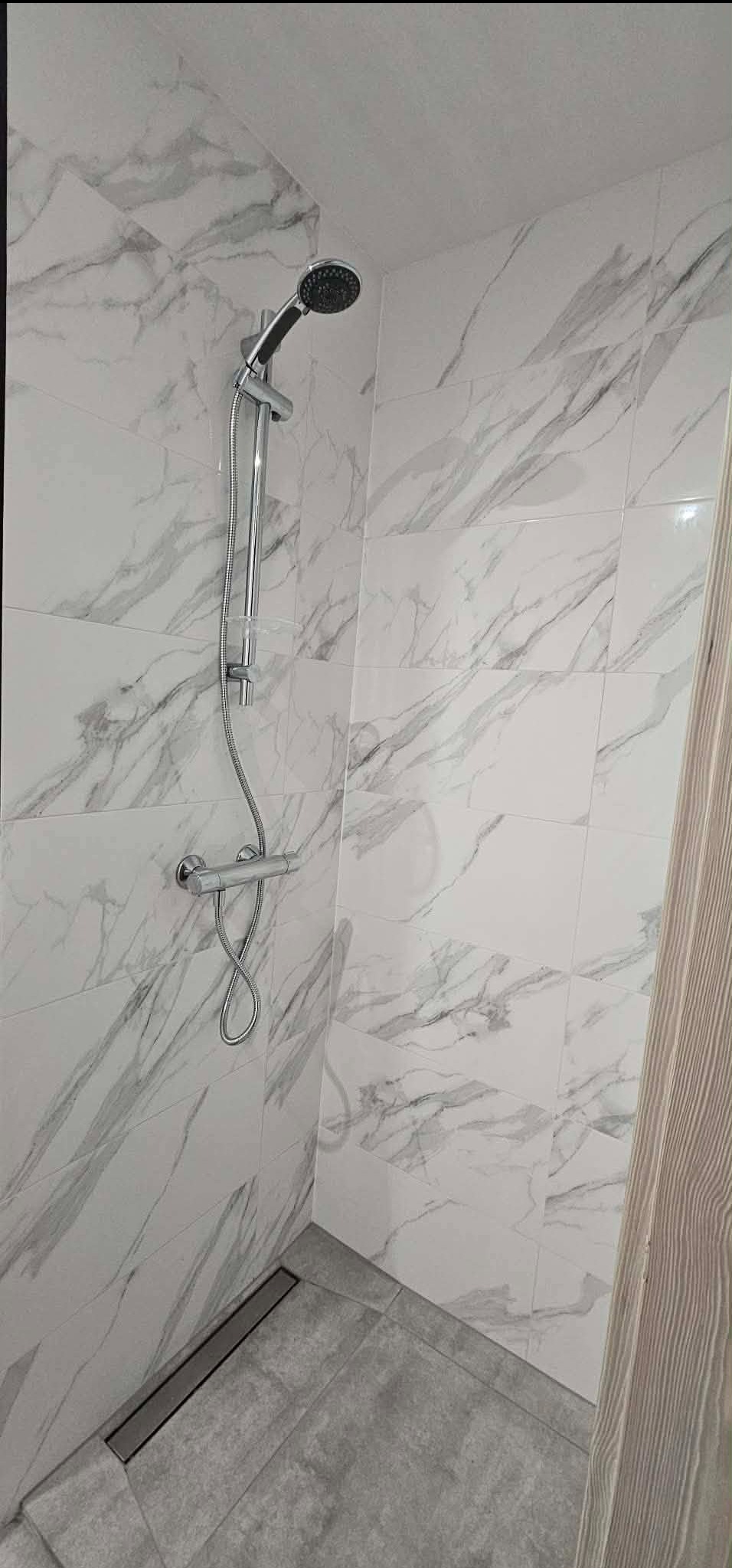 Kabina prysznicowa z odpływem liniowym i baterią termostatyczną, wykończona białymi płytkami imitującymi marmur. Minimalistyczny design.