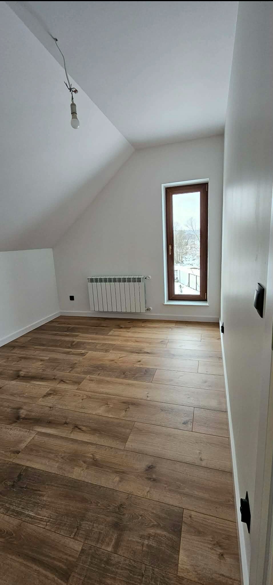 Jasne poddasze z drewnopodobną podłogą, białymi ścianami i oknem z widokiem na zimowy krajobraz. Wisząca żarówka dodaje surowego charakteru.