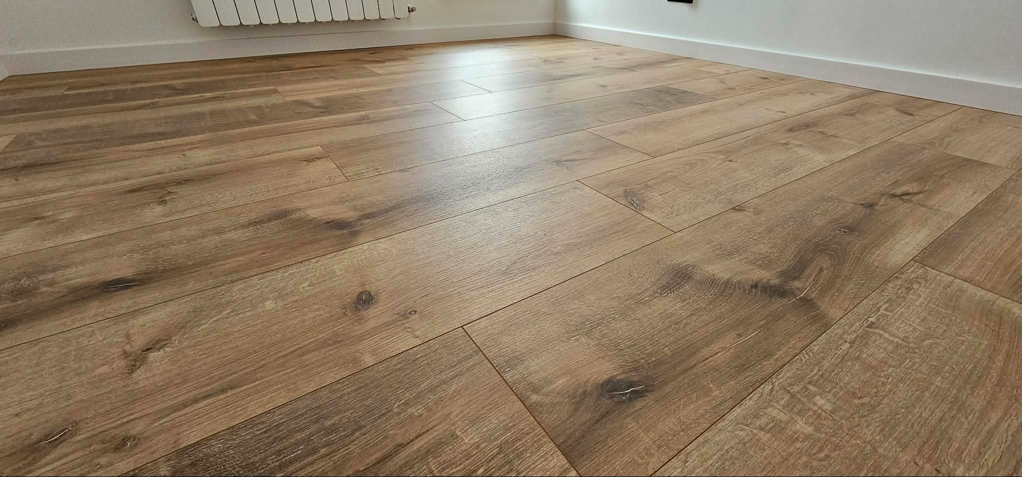 Ułożona podłoga z paneli w odcieniach brązu, widok z niskiej perspektywy, ukazujący strukturę drewna i łączenia. W tle biała listwa przypodłogowa i fragment grzejnika.