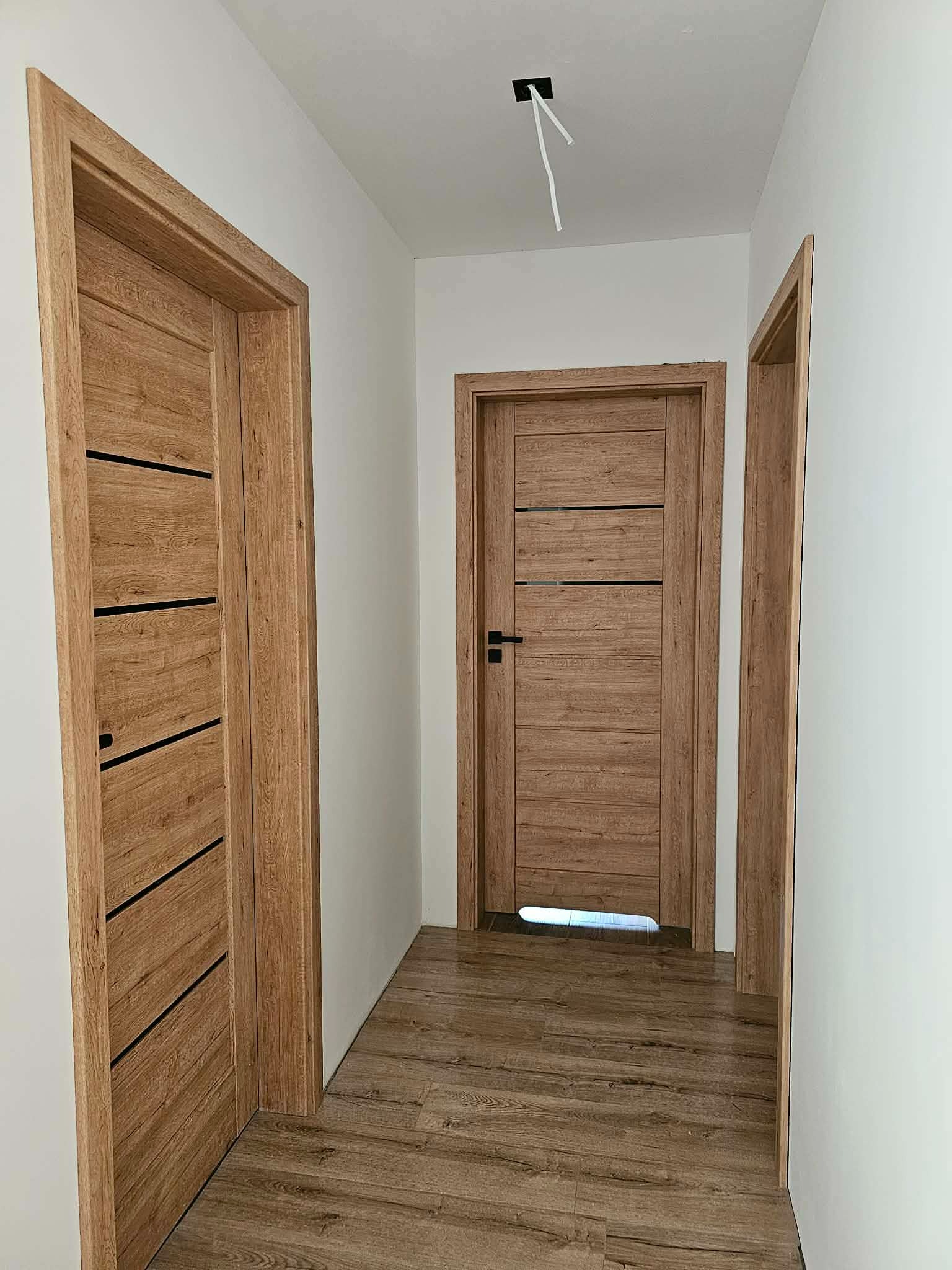 Nowoczesne, drewniane drzwi wewnętrzne z czarnymi akcentami, zamontowane w jasnym korytarzu z panelami podłogowymi. Widoczne kable wystające z sufitu.