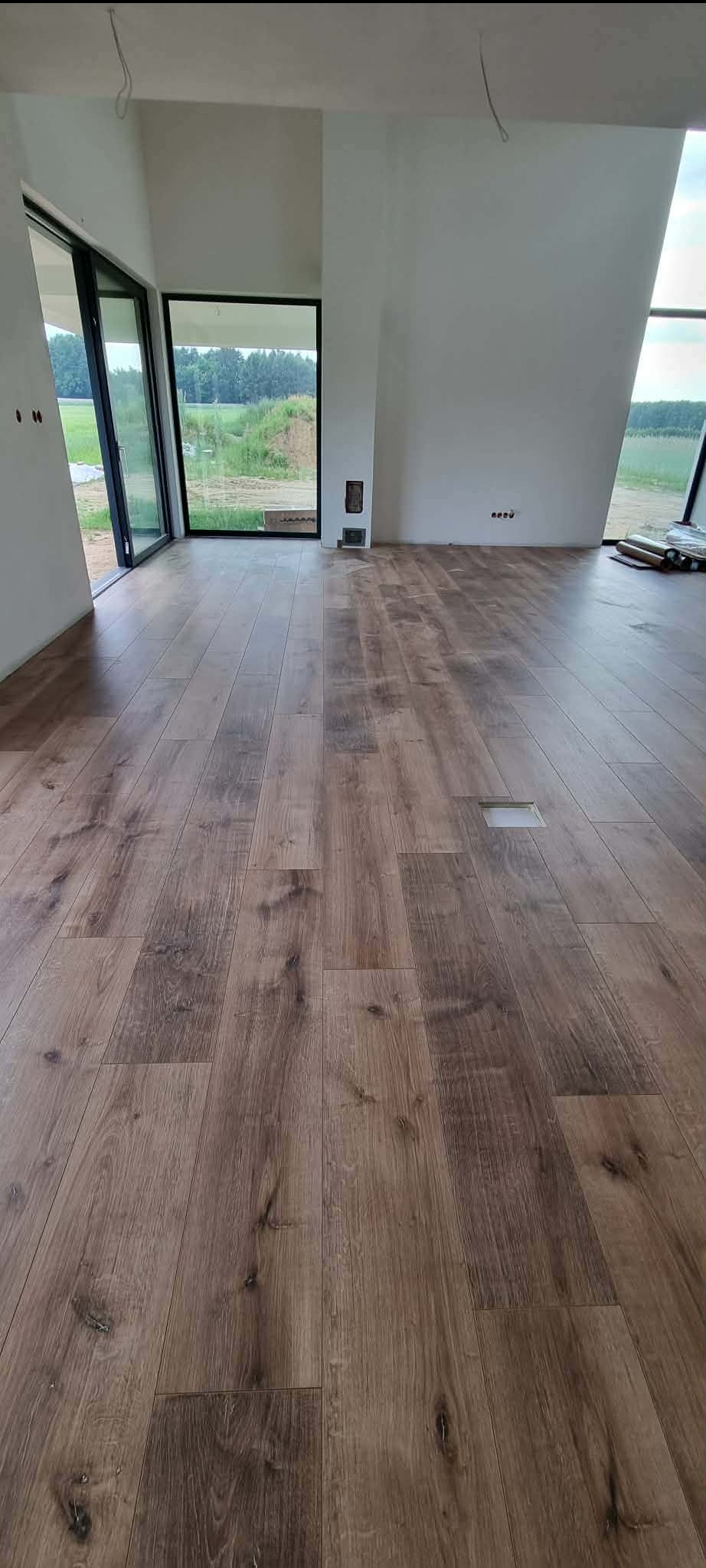 Ułożona podłoga z paneli w jasnym wnętrzu z dużymi oknami. Widoczny fragment niezakończonej instalacji podłogowej. Naturalne światło dzienne.
