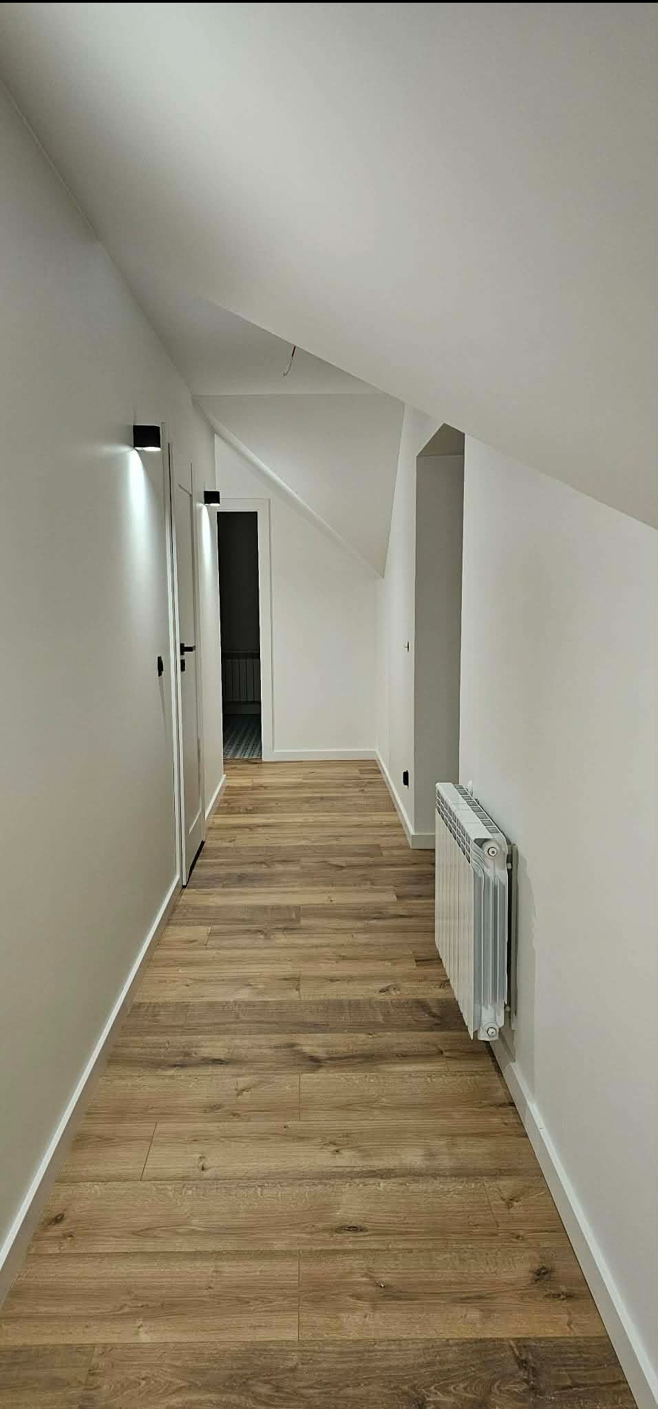 Biały korytarz na poddaszu z drewnianą podłogą, grzejnikiem i czarnymi lampami ściennymi. Widoczne drzwi i fragment schodów.