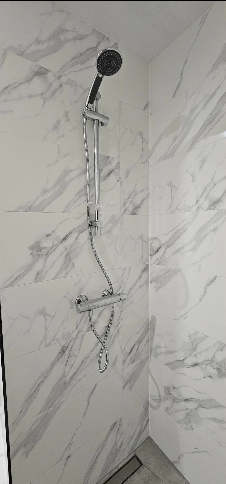 Nowoczesna kabina prysznicowa z marmurowymi płytkami, chromowaną armaturą i odpływem liniowym. Minimalistyczny design, jasne wnętrze.