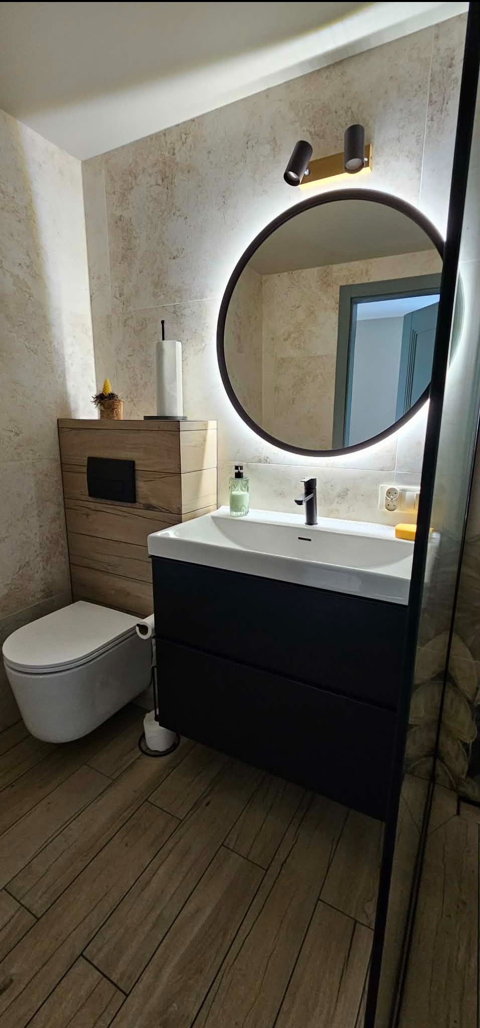 Nowoczesna łazienka z podświetlanym lustrem, drewnianą szafką podtynkową i czarną umywalką. Uwagę przykuwają płytki imitujące drewno na podłodze i ścianach.