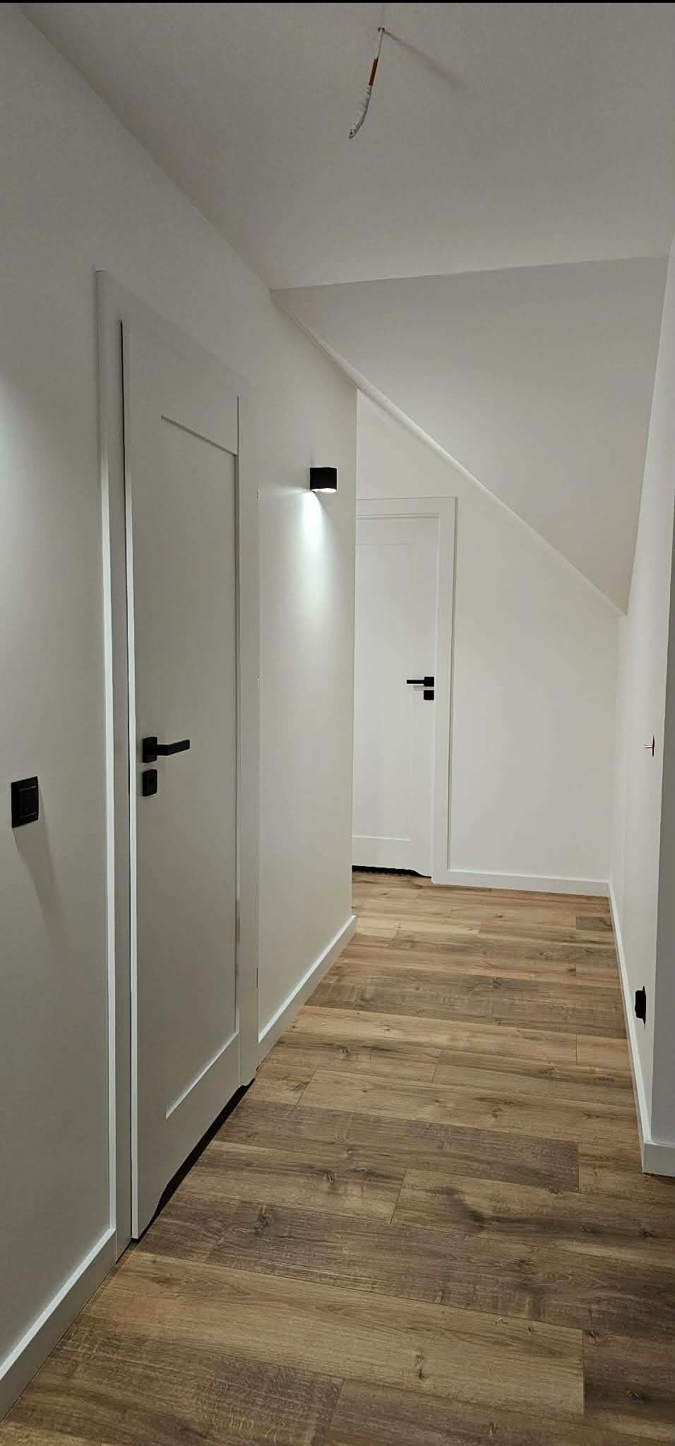 Biały korytarz z drewnopodobną podłogą, oświetleniem punktowym i minimalistycznymi drzwiami z czarnymi klamkami. Widok na skos dachu i kolejne drzwi w głębi.