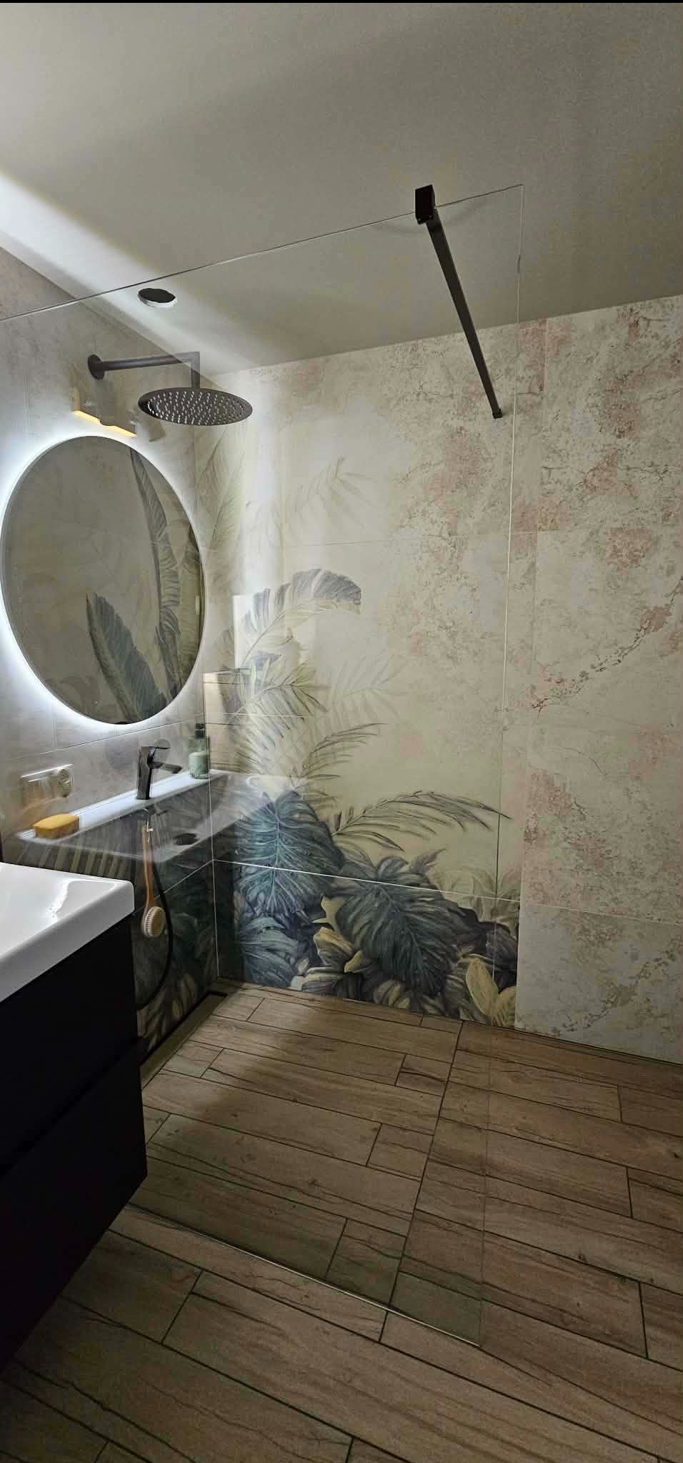 Nowoczesna łazienka z kabiną typu walk-in, płytkami z motywem roślinnym i okrągłym lustrem z podświetleniem LED. Drewnopodobna podłoga dodaje ciepła wnętrzu.