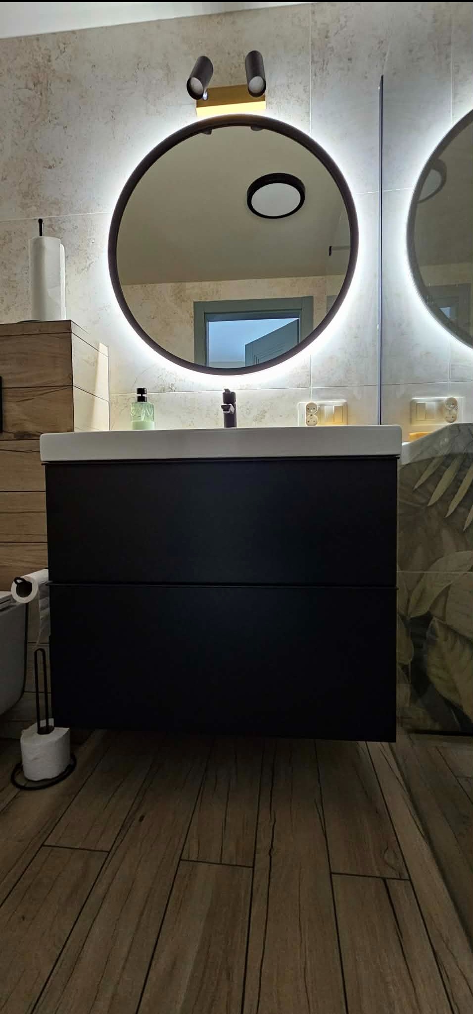 Nowoczesna łazienka z czarną szafką pod umywalką, okrągłym lustrem z podświetleniem LED i drewnopodobną podłogą. Widoczne detale wykończenia.