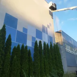 Mycie elewacji budynku z geometrycznym wzorem z podnośnika koszowego, widoczne smugi czystej elewacji, rząd iglaków u dołu kadru i błękitne niebo.