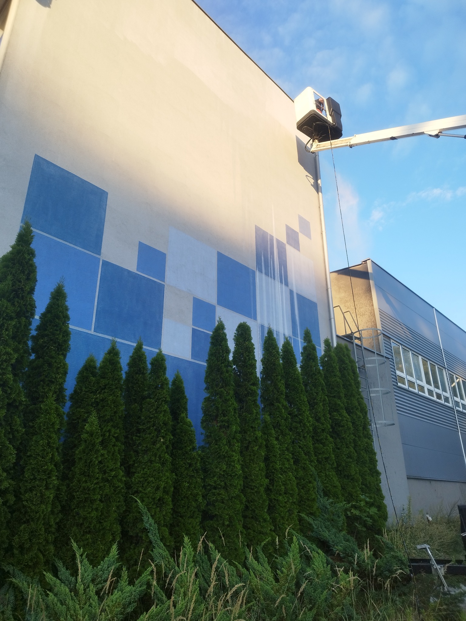 Mycie elewacji budynku z geometrycznym wzorem z podnośnika koszowego, widoczne smugi czystej elewacji, rząd iglaków u dołu kadru i błękitne niebo.