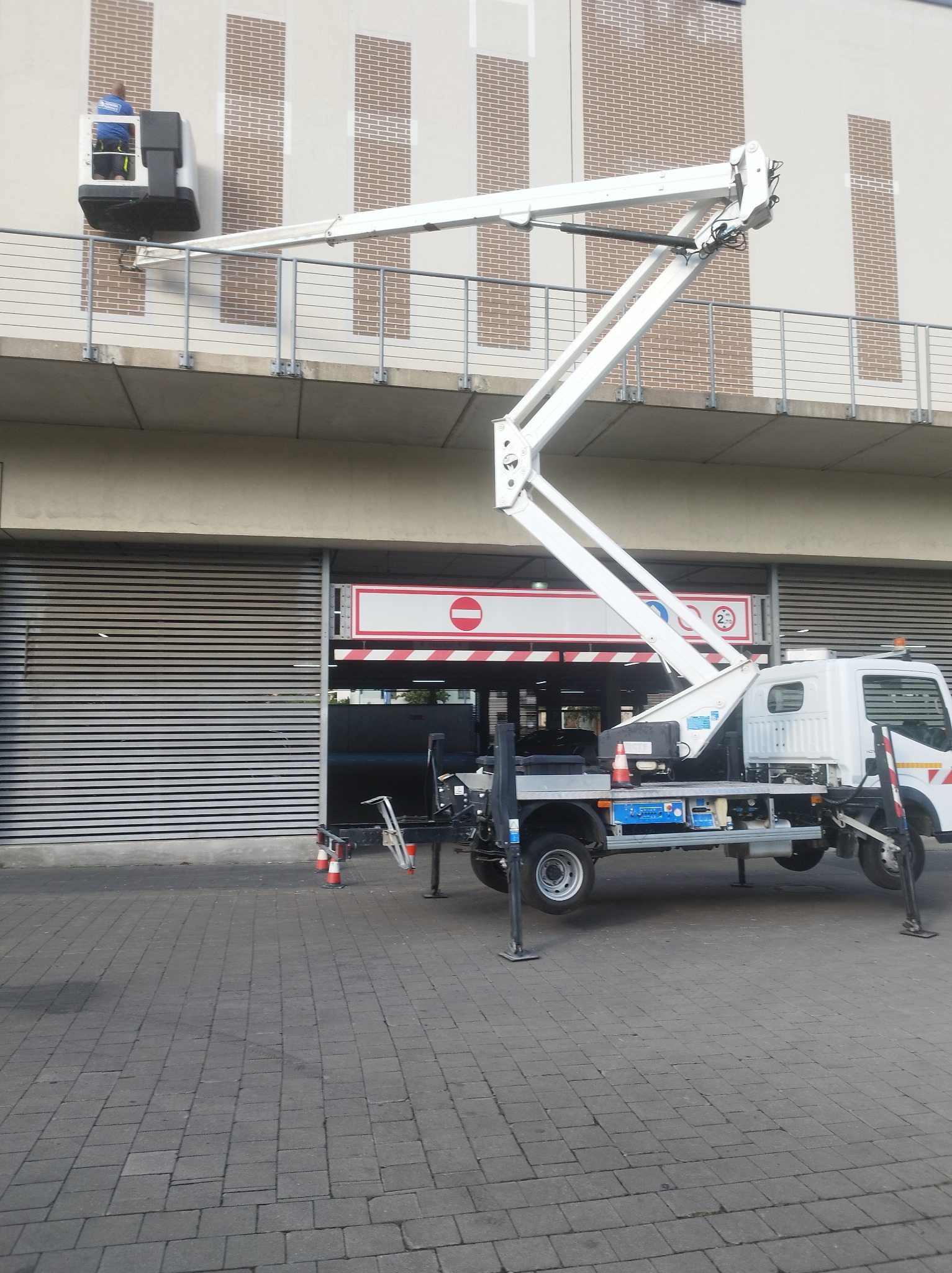 Biały podnośnik koszowy na tle budynku z szarymi panelami i ceglanymi wstawkami. W koszu widoczna osoba w niebieskiej koszulce. Widoczne barierki i zaparkowany samochód.