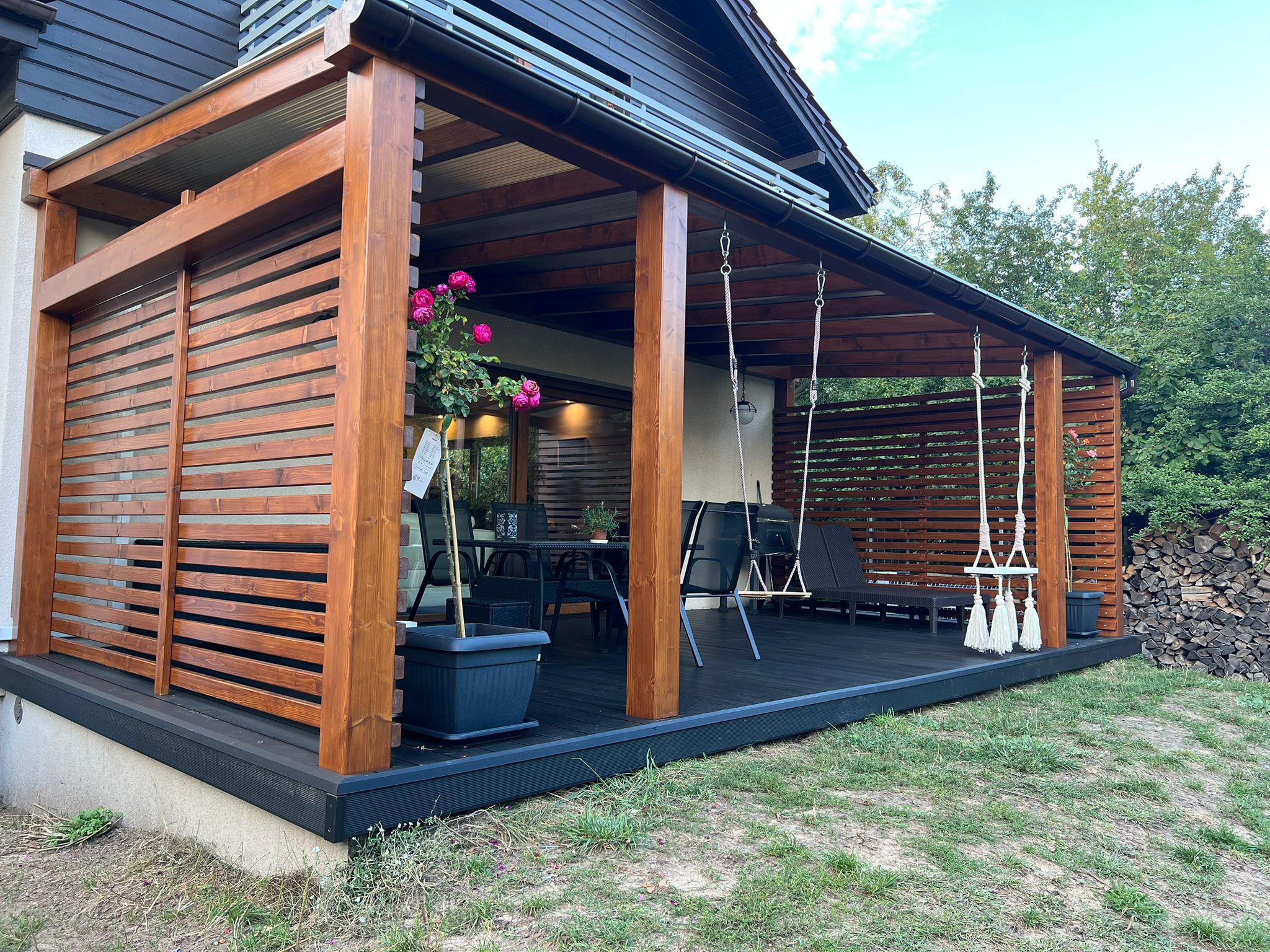Zadaszony taras z drewnianą pergolą i czarną podłogą kompozytową. Na tarasie meble ogrodowe, huśtawka i donica z kwiatami. Widoczne drewniane panele przesłaniające.