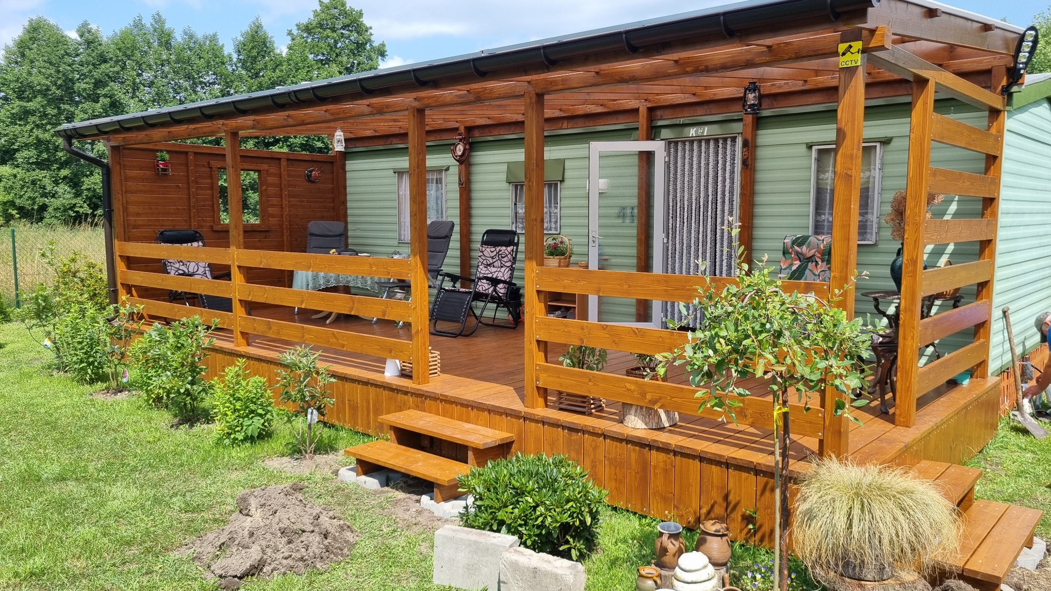 Drewniana zabudowa tarasu z balustradą, zadaszeniem i schodkami. Na tarasie meble ogrodowe, a przed nim trawnik z roślinami. Widoczny numer domu K-1 i lampa.
