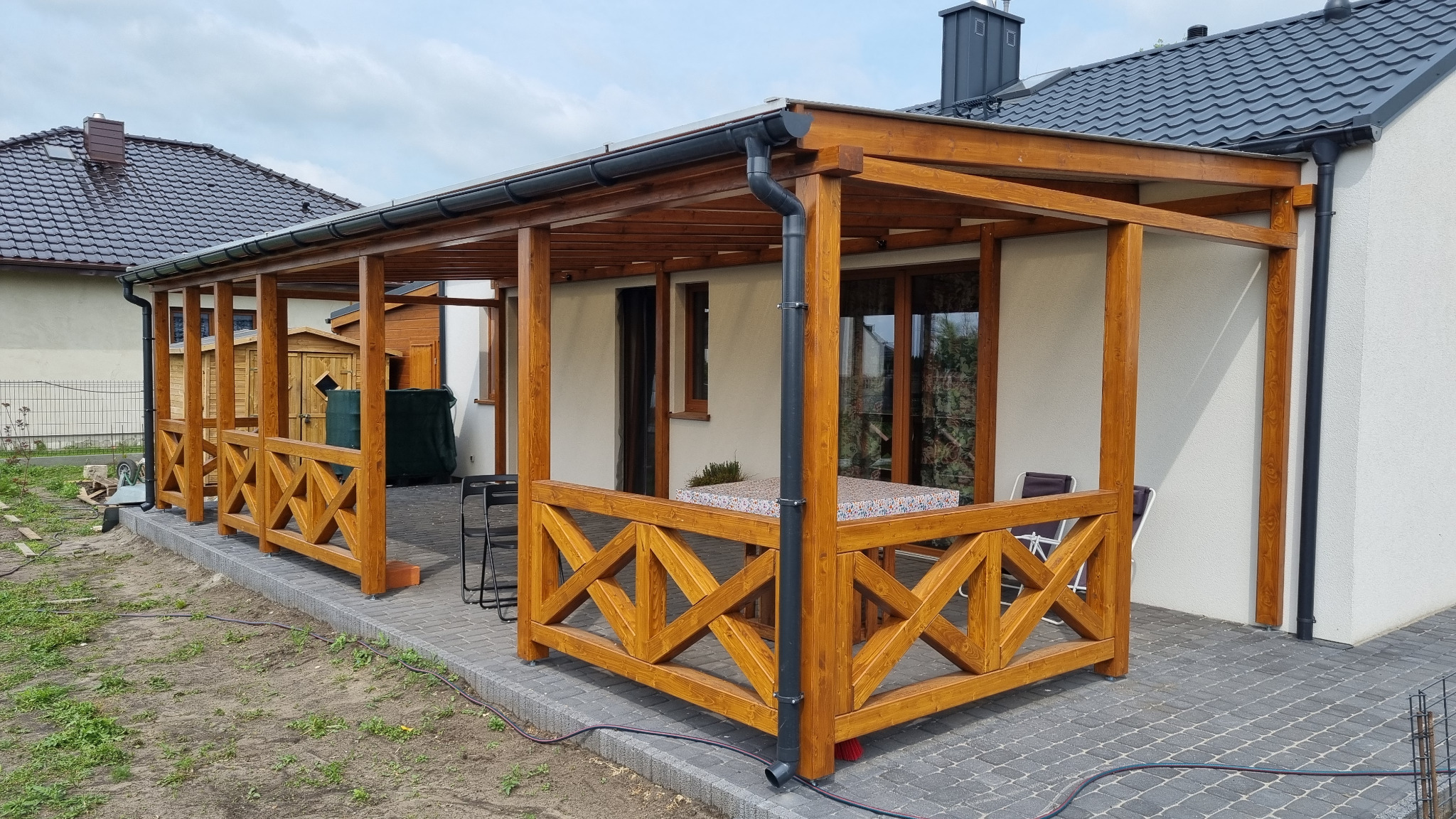 Drewniana pergola z ozdobną balustradą, przylegająca do domu z szarą dachówką. Widoczne meble ogrodowe i kostka brukowa na podłożu.