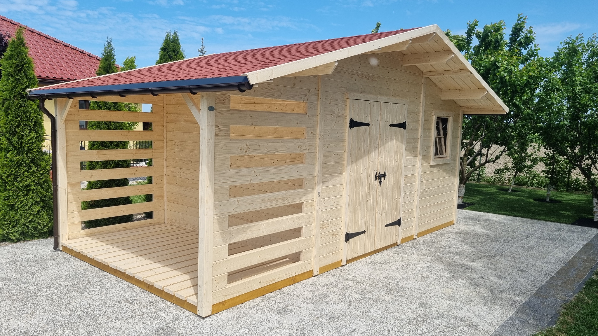 Drewniana altana z czerwonym dachem, otwartą przestrzenią z boku i zamkniętą częścią z drzwiami, umieszczona na kostce brukowej w ogrodzie.