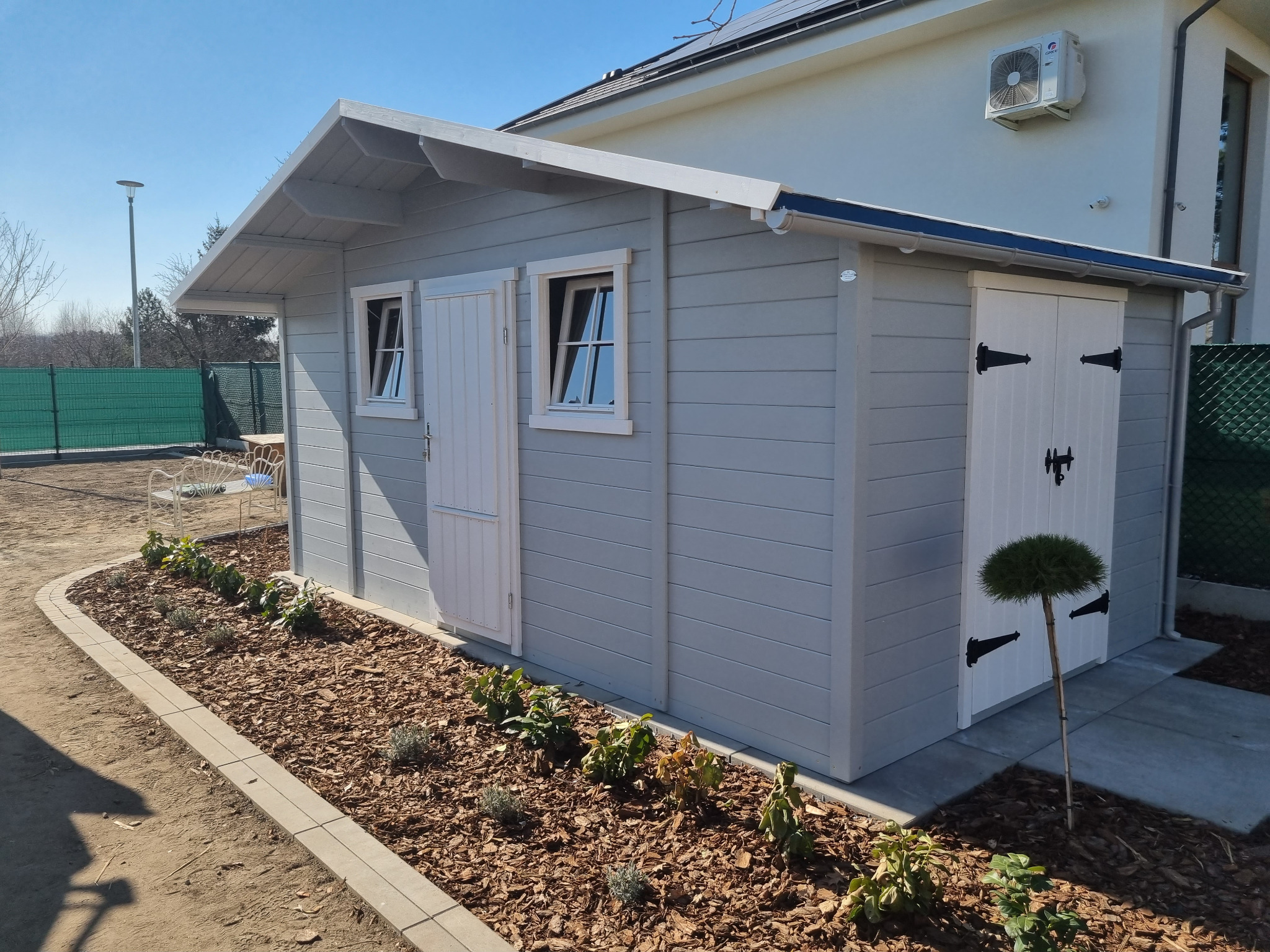 Szara, drewniana altana z białymi drzwiami i oknami, otoczona rabatą z korą i niskimi roślinami, na tle domu z klimatyzacją i zielonego ogrodzenia.