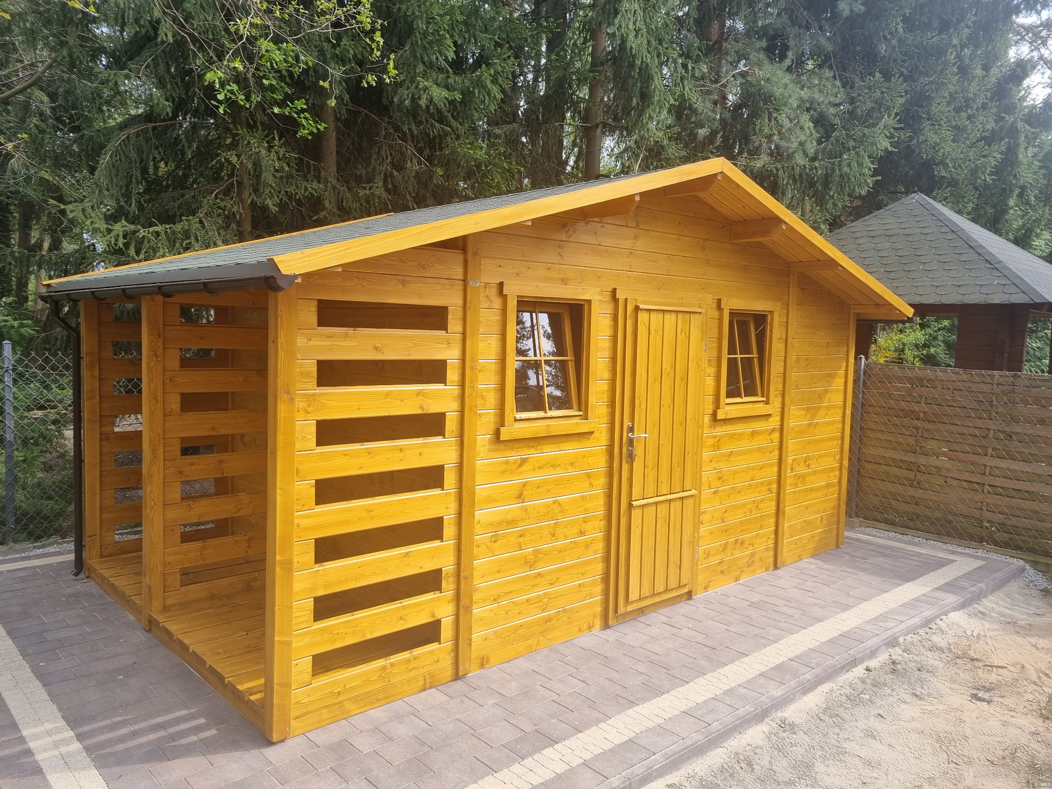 Żółta, drewniana altana z oknami i drzwiami, zadaszona, z otwartą przestrzenią z boku, na kostce brukowej, w tle drzewa i fragment innej altany.