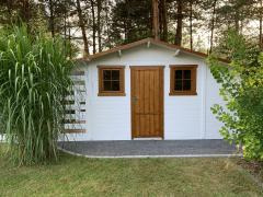 Biała altana z brązowymi ramami okien i drzwiami, otoczona zielenią trawy i drzew, z widocznymi schodkami z boku. Ujęcie w naturalnym oświetleniu.