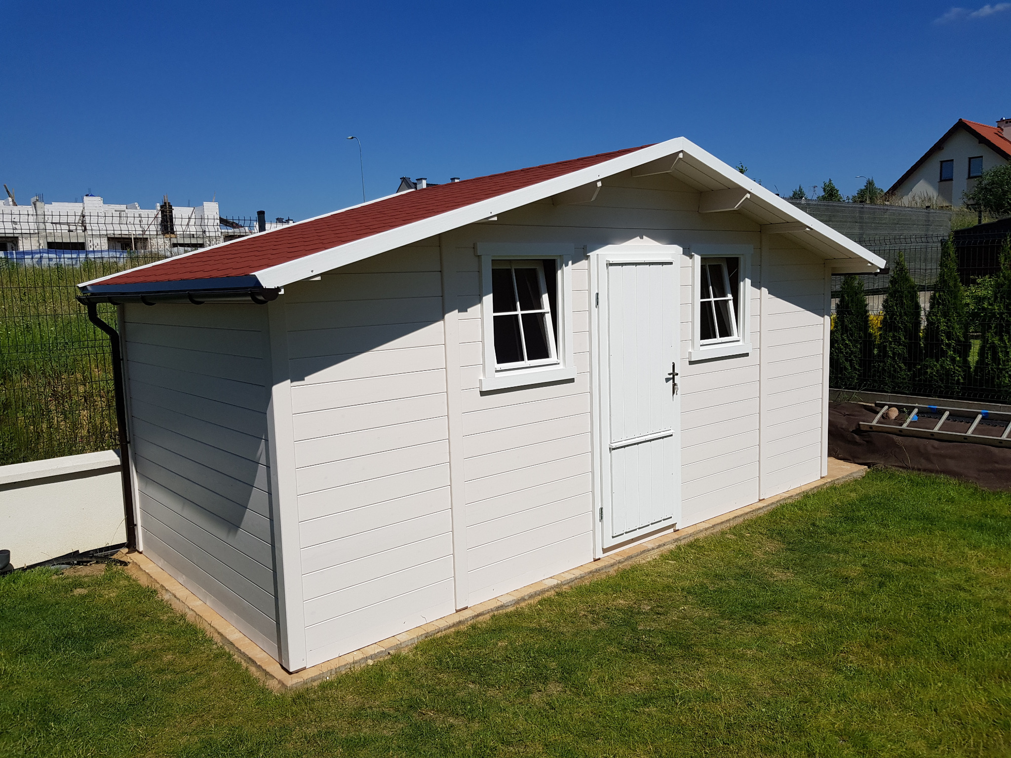 Biała, drewniana altana z czerwonym dachem na trawniku w słoneczny dzień. Widoczne okna i drzwi altany, obok trawa i elementy otoczenia.