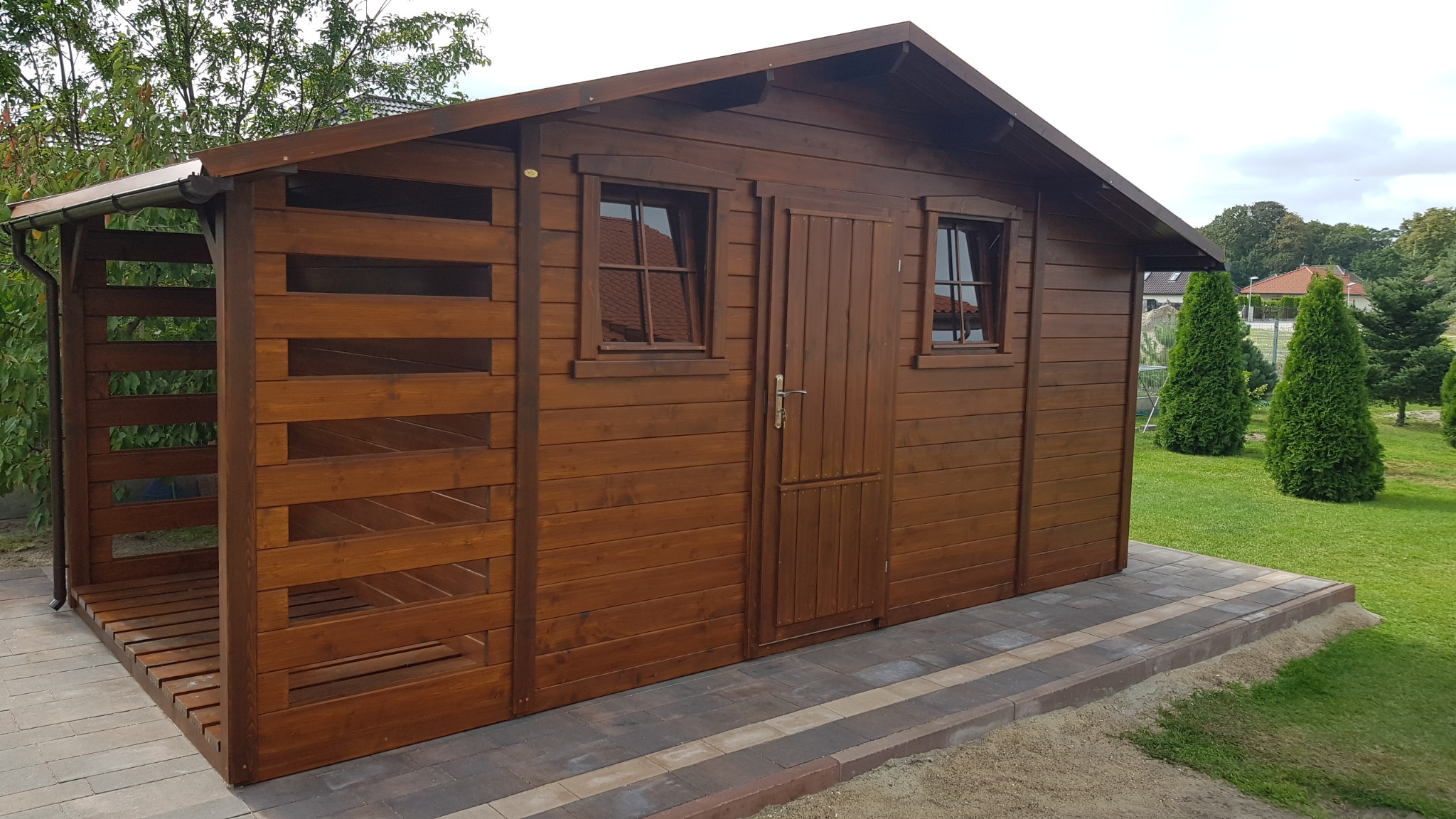 Ciemnobrązowa, drewniana altana z oknami i drzwiami, ustawiona na kostce brukowej. Widoczne elementy konstrukcyjne i otoczenie z zielenią.